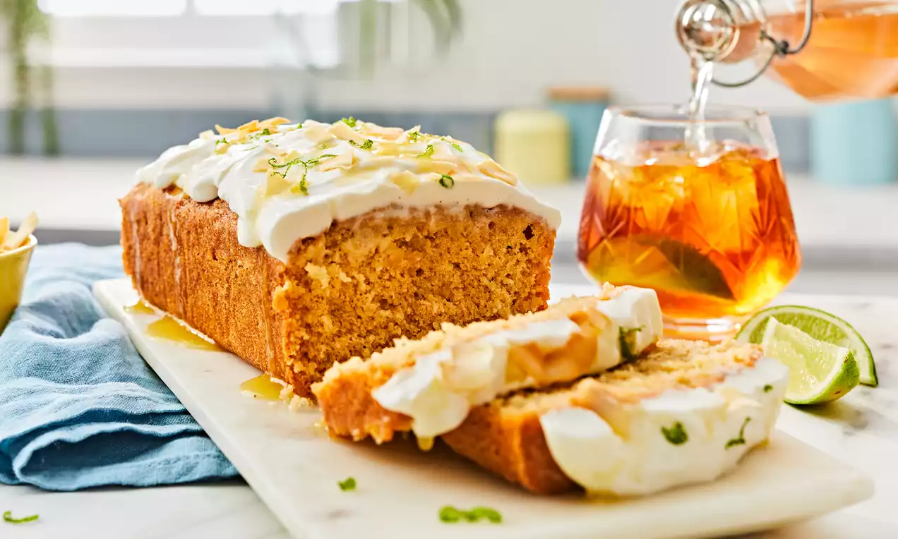 Golden loaf-shaped rum cake with moist crumb, topped with smooth white icing, lime zest, and syrup on a marble board