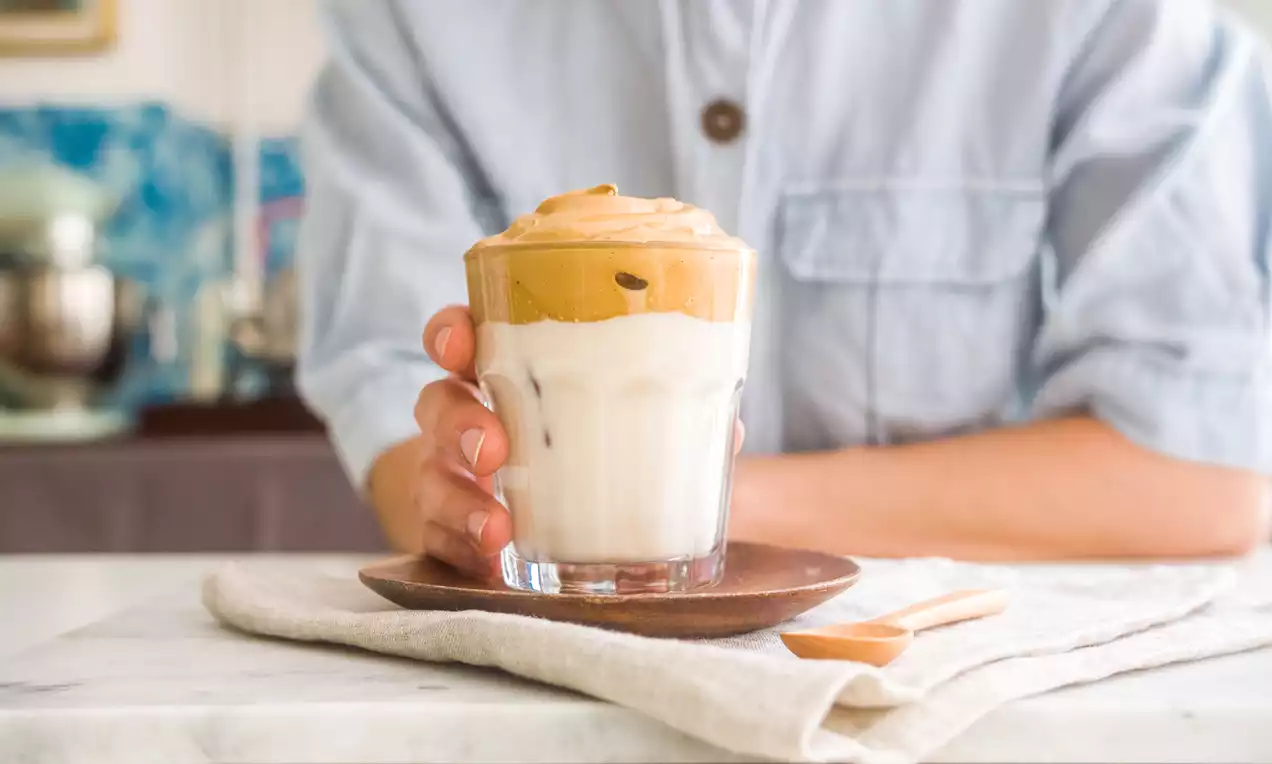 Café Dalgona con espuma dorada y cremosa sobre leche blanca en vaso transparente, en un plato marrón sobre tela beige