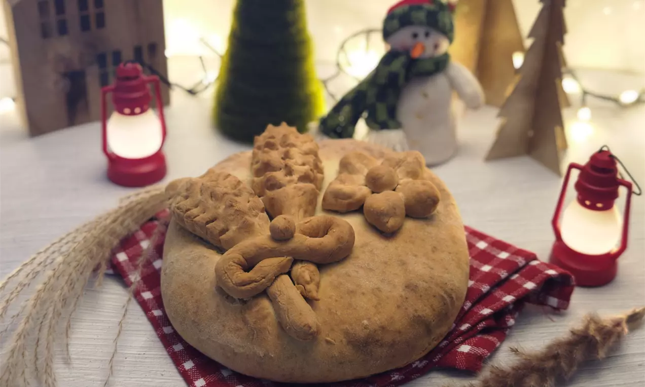 Кръгла содена питка с равномерно златиста коричка, украсена с тестени орнаменти върху червено каре на бяла маса сред празнична украса