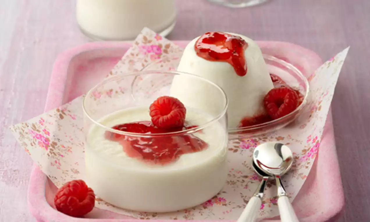 Panna cotta blanca y lisa servida en vaso y desmoldada, con coulis rojo brillante y frambuesas frescas sobre bandeja decorativa rosa