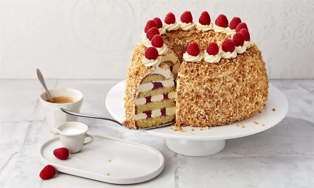 Кръгъл сладкиш с златиста текстура от препечени ядки, кремово покритие и малини в горния край, разрязан със слоеве крем и плодове