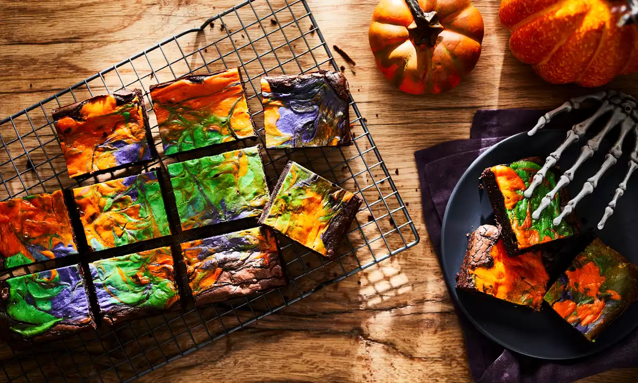 Square chocolate brownies with vibrant swirls of orange, green, and purple icing, arranged on a cooling rack and dark plate near pumpkins