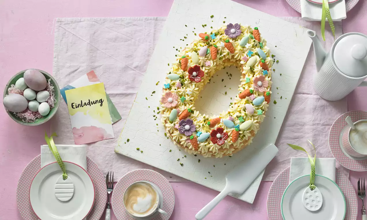 Ovaler Kuchen mit heller Creme und bunten Zuckerdekoren, umgeben von pastellfarbenem Geschirr und einer Einladung auf rosafarbenem Tischtuch