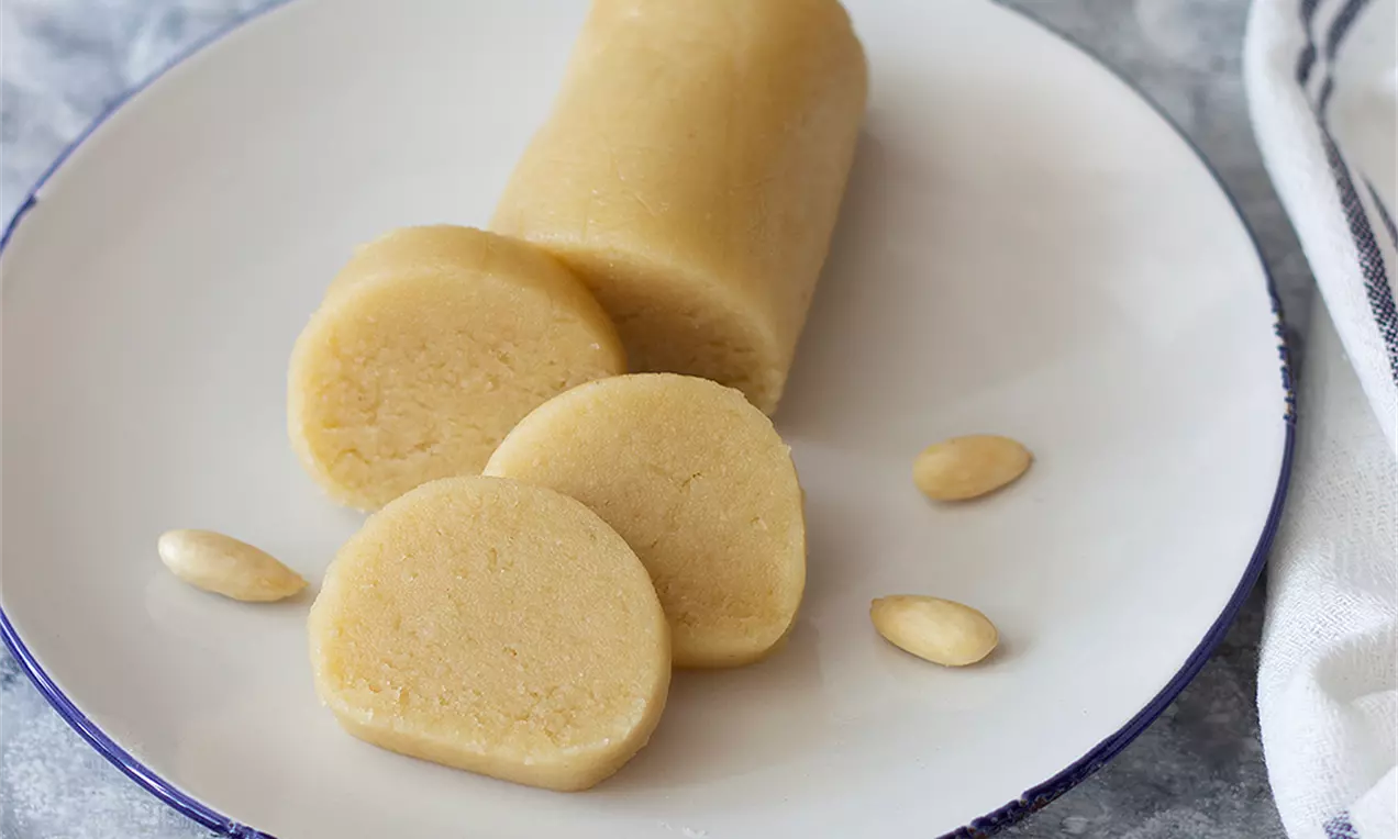 Светложълт бадемов марципан в цилиндрична форма, нарязан на гладки дебели кръгове върху бяла чиния, с няколко цели белени бадема