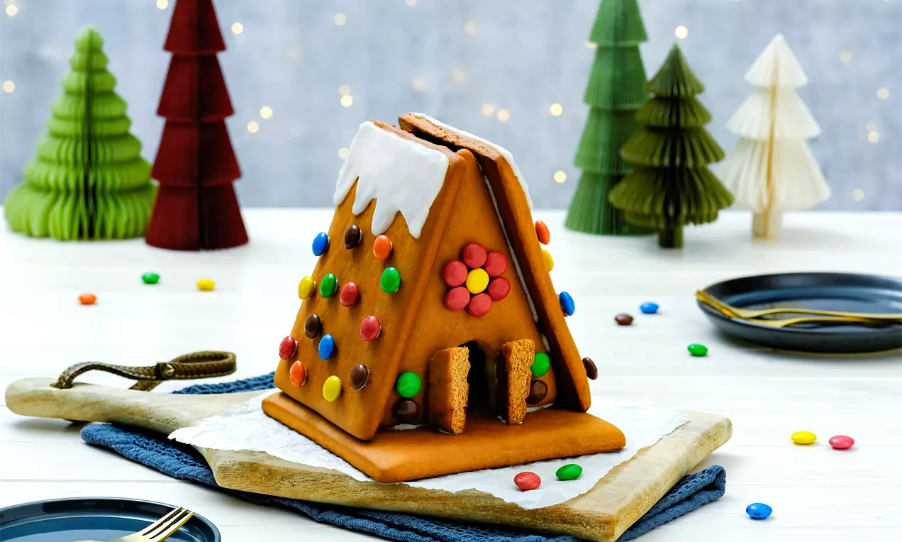 Къщичка от кафяво медено тесто с бяла глазура по покрива и разноцветни бонбони, поставена върху дървена дъска с фон от коледни елхи