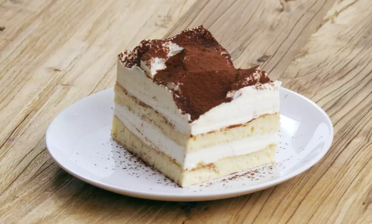 Een vierkante plak tiramisu met lagen luchtige mascarponecrème en cake, bestrooid met cacaopoeder, op een wit bord op een houten tafel