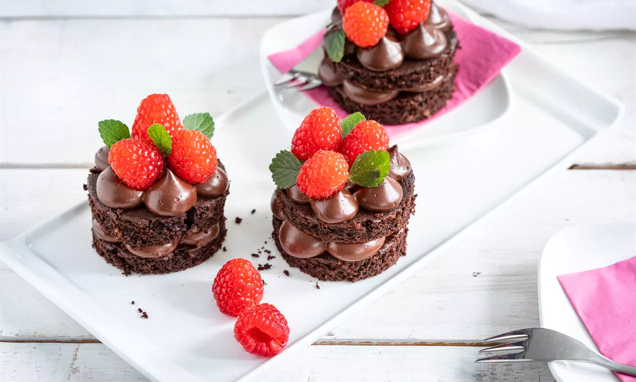 Шоколадови пластове с крем, подредени на кръгли порции с свежи малини и листа мента върху бяла правоъгълна чиния