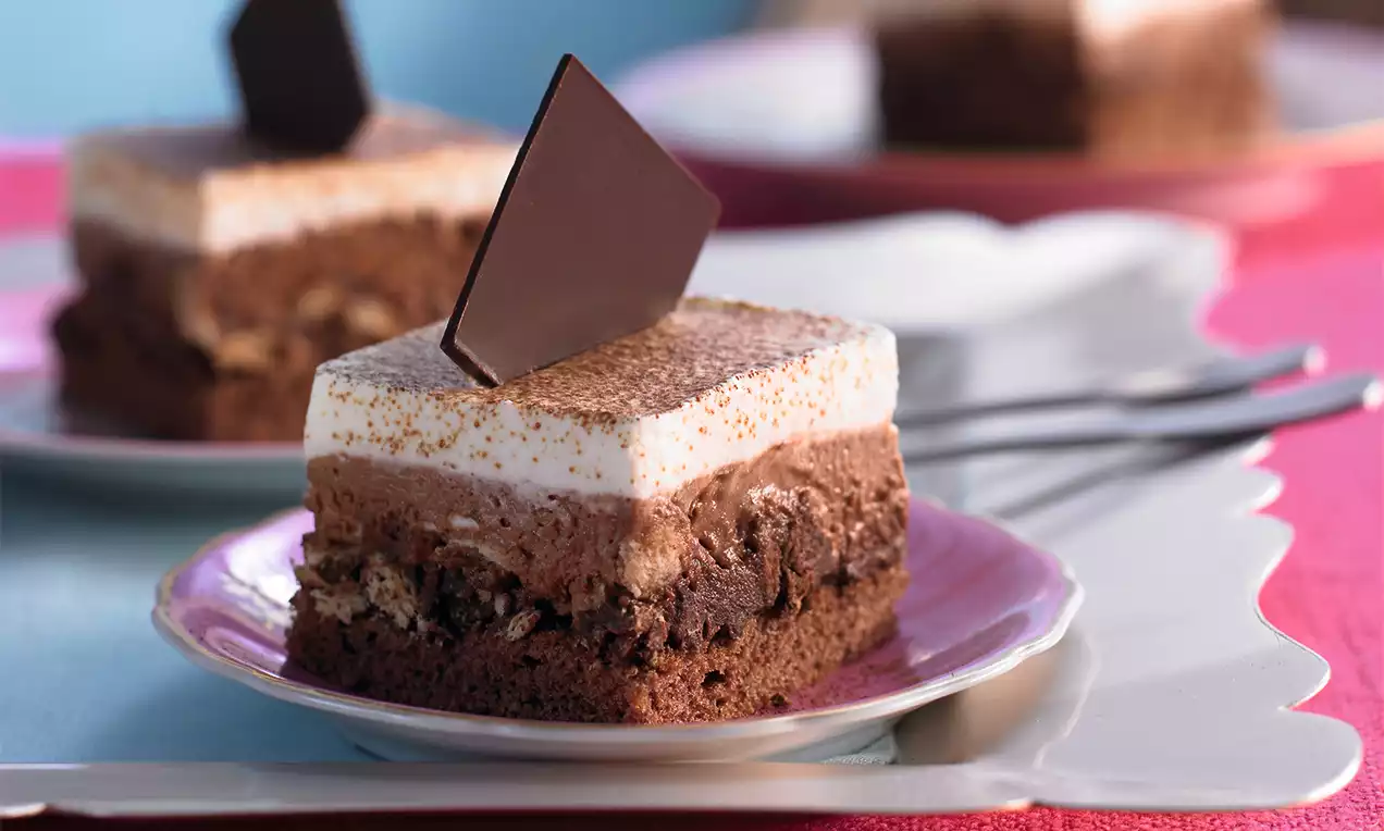 Čtvercové vrstvy čokoládového korpusu, krému a šlehačky, zasypané kakaovým práškem, ozdobené čokoládovým trojúhelníkem na růžovém talíři