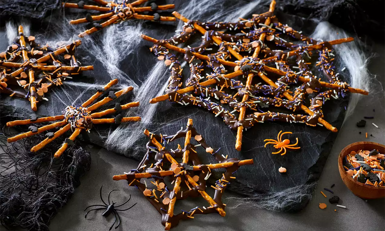 Sprøde edderkoppespind af saltstænger med mørk chokolade og farvet krymmel på sort skiferplade med kunstige edderkopper og dekoreret baggrund