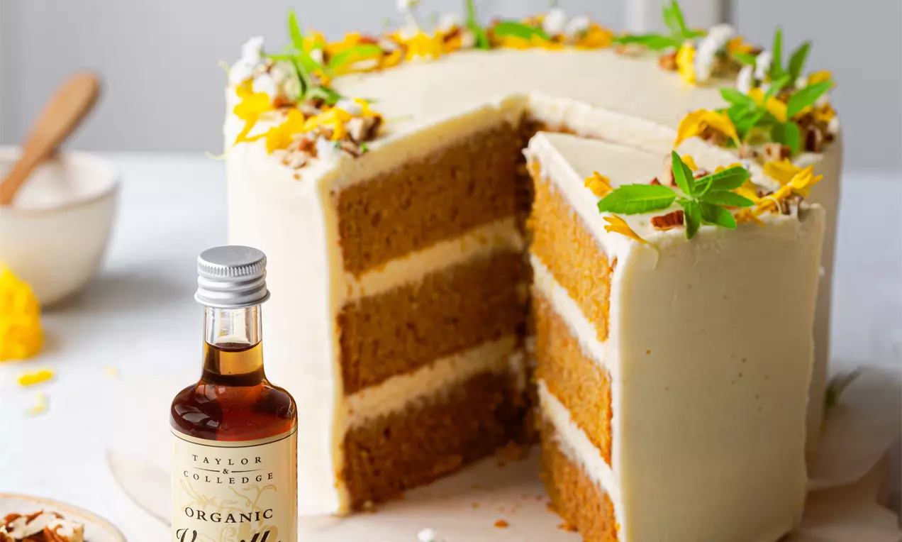 Kolmikerroksinen porkkanakakku pehmeällä vaalealla kuorrutteella, koristeltu keltaisilla kukilla ja vihreillä mintunlehdillä vaalealla tarjoilualustalla