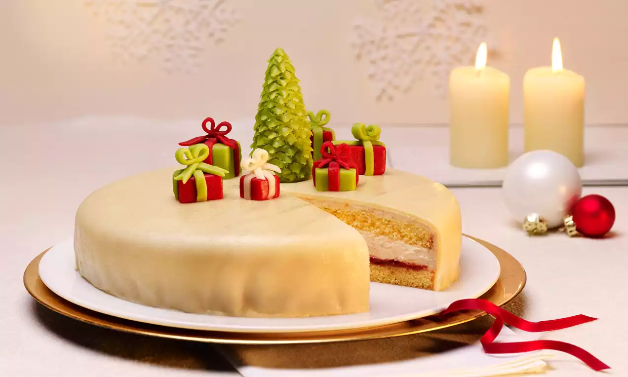 Rund kake med glatt beige marsipantrekk, et utskåret stykke som viser lag med lys kakebunn og fyll, dekorert med små marsipanpakker og et juletre på en hvit tallerken med rødt bånd