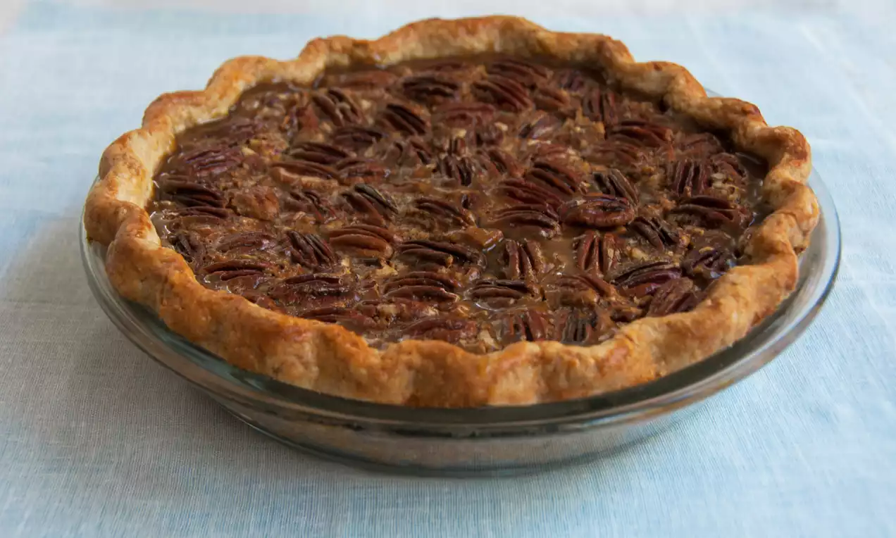 Tarte aux pacanes dorées avec une croûte légèrement ondulée, garnie de noix brillantes et disposée dans un plat en verre transparent sur un tissu bleu