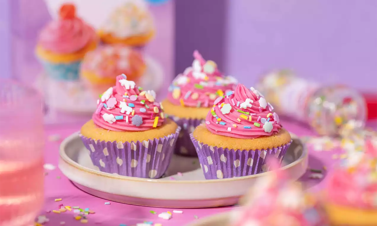 Cupcakes met roze crème en kleurrijke strooisels in paarse bakvormpjes met stippen op een licht bord tegen een paarse achtergrond