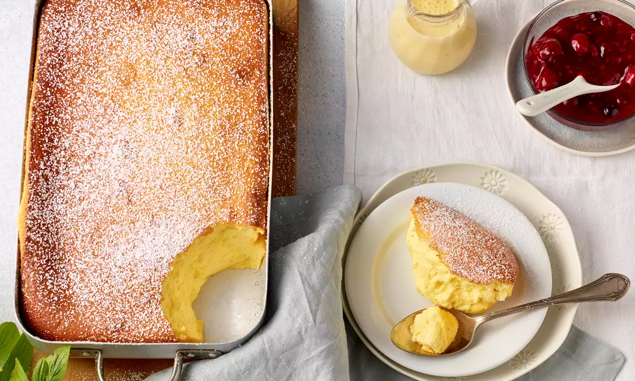 Topfenauflauf mit goldbrauner Kruste auf weißem Teller, dazu leuchtend rote Kirschgrütze, samtig helle Vanillesoße und Puderzucker