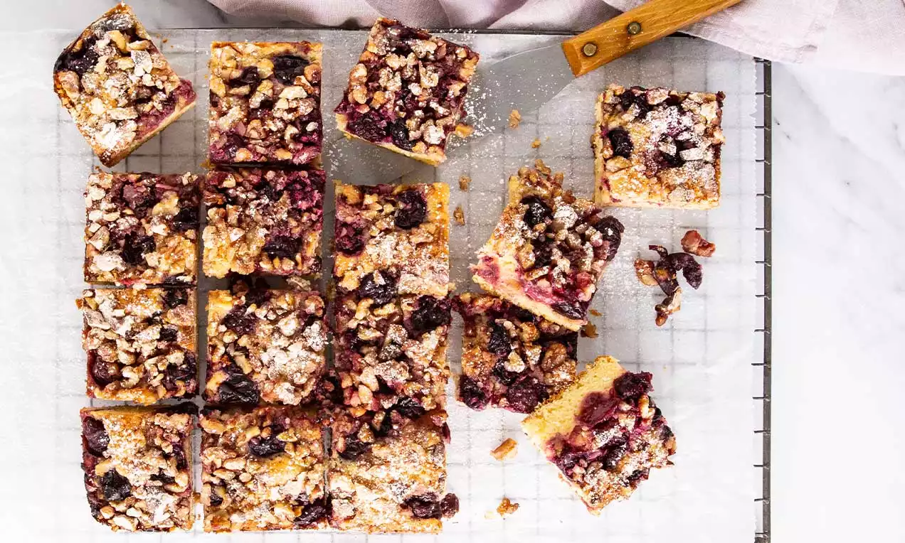 Carrés dorés de gâteau moelleux parsemés de cerises juteuses et éclats de noix saupoudrés de sucre glace sur une grille blanche