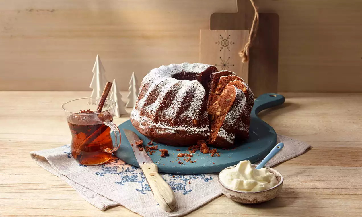 Lebkuchengugelhupf mit dunkler Kruste und Puderzucker, angeschnitten auf blauem Brett mit Rahm und Tee, umgeben von weihnachtlicher Deko