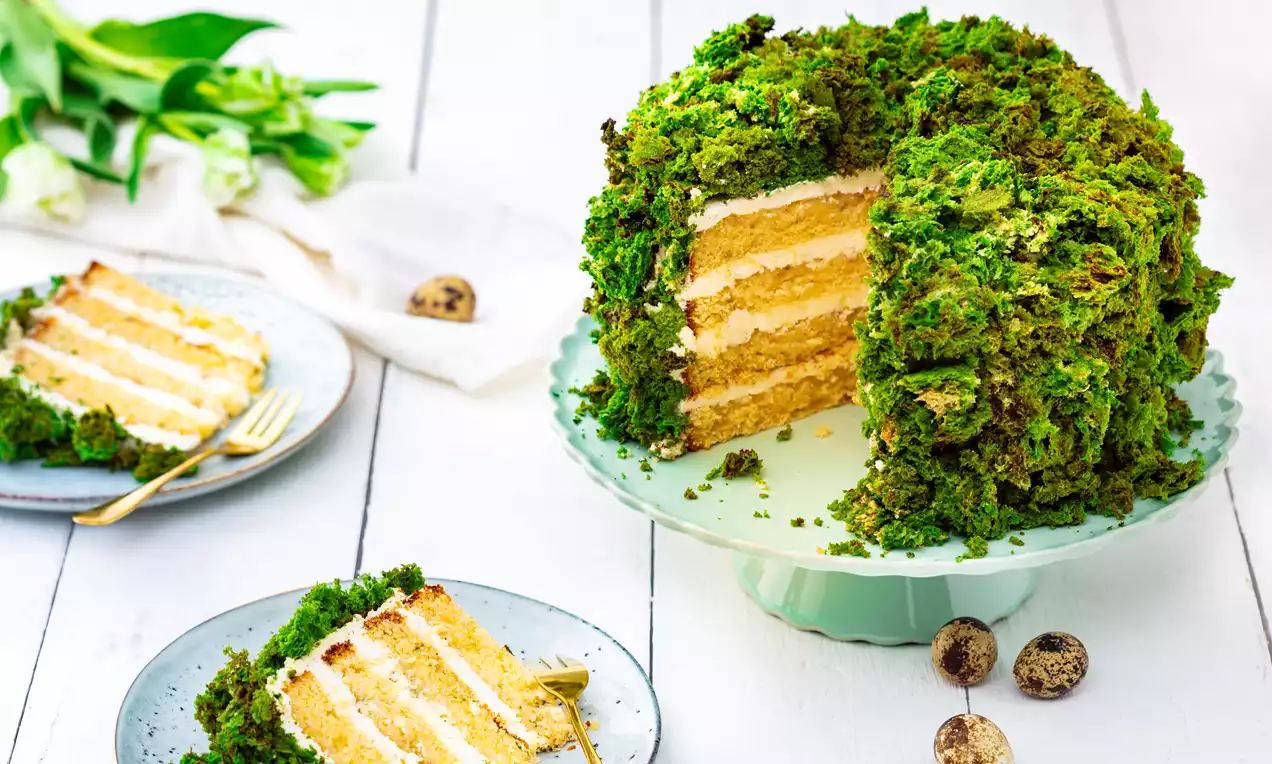 Høy, rund kake med lyse lag av krem og kakebunn dekket med løs, grønn smuldredekor på lyseblått kakefat med utskårede stykker på asjetter