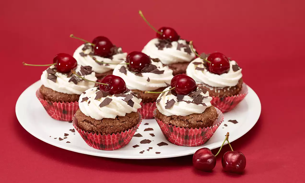 Cupcakes au chocolat avec crème blanche, copeaux de chocolat noir et cerises brillantes sur des caissettes rouges, disposés sur une assiette blanche