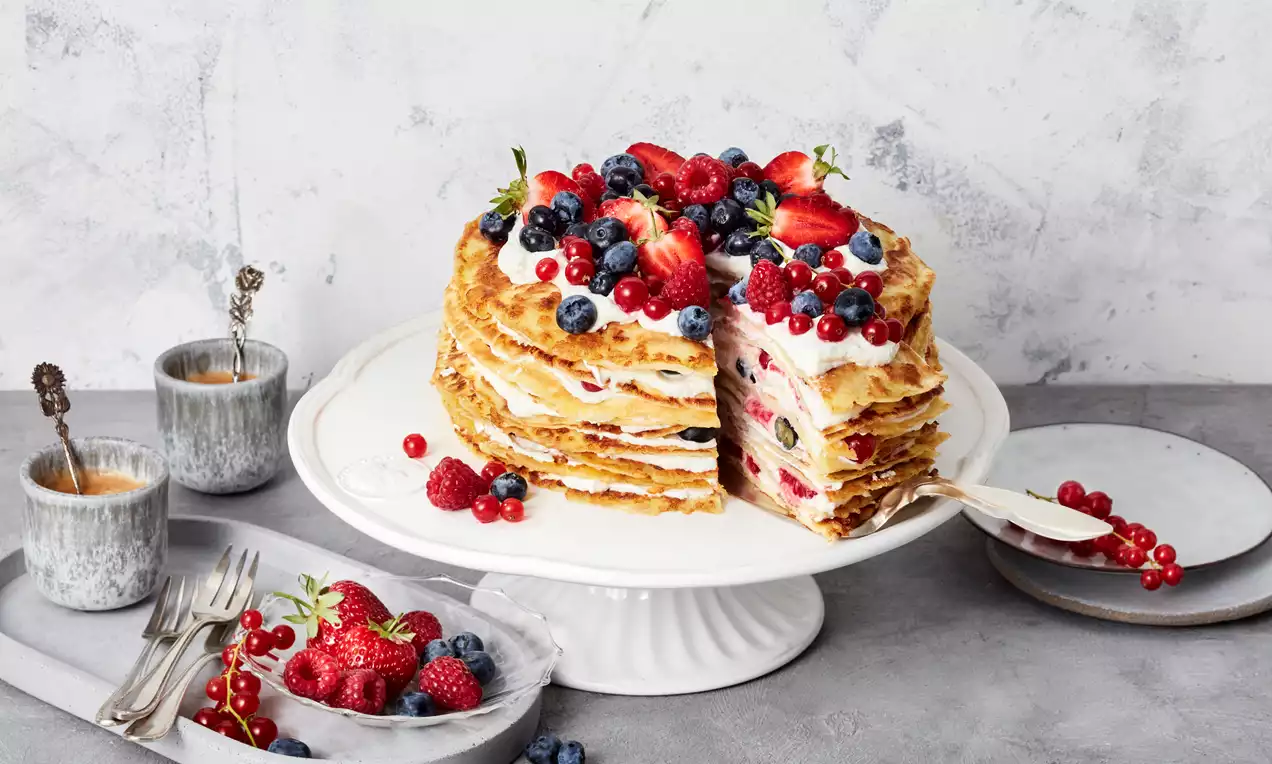 Pfannkuchentorte aus dünnen goldbraunen Schichten, gefüllt mit heller Creme und Beeren, garniert mit frischen Früchten auf weißer Tortenplatte