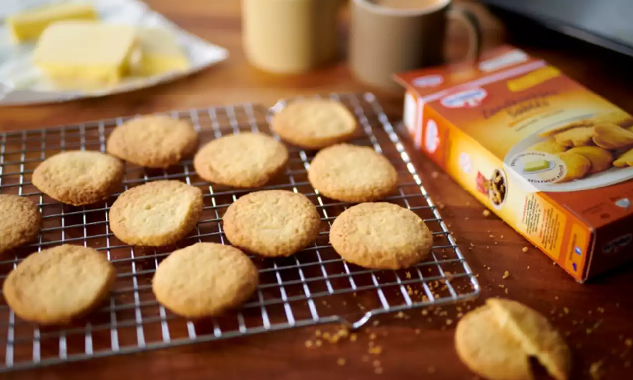 Zandkoekjes goudbruin en rond met kruimelige textuur op een metalen koelrek, naast een verpakking, kopjes koffie en een botervlootje