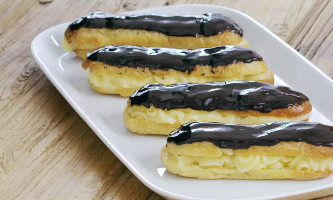 Licht goudbruine eclairs gevuld met romige gele vulling en bedekt met glanzend donkere chocoladelaag op een wit bord