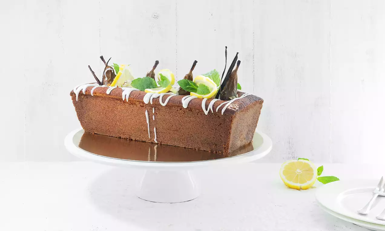 Rechthoekige, goudbruine gin-toniccake met glanzend witte glazuurstrepen, partjes peer, muntblaadjes en limoen op een witte taartstandaard