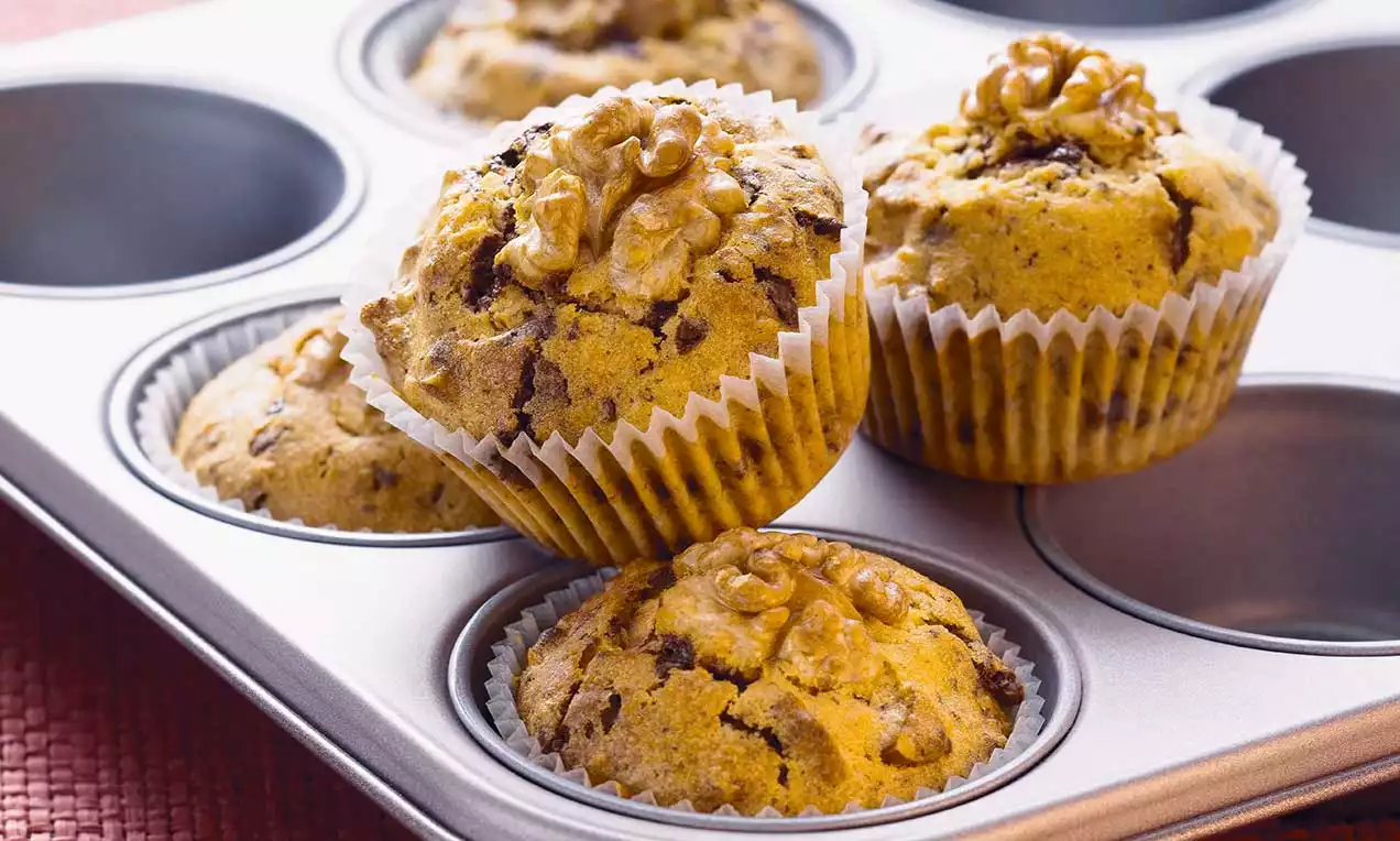 Saftige, goldbraune Muffins mit schokoladigen Einsprengseln in Papierförmchen, garniert mit Walnüssen und auf einem Muffinblech arrangiert