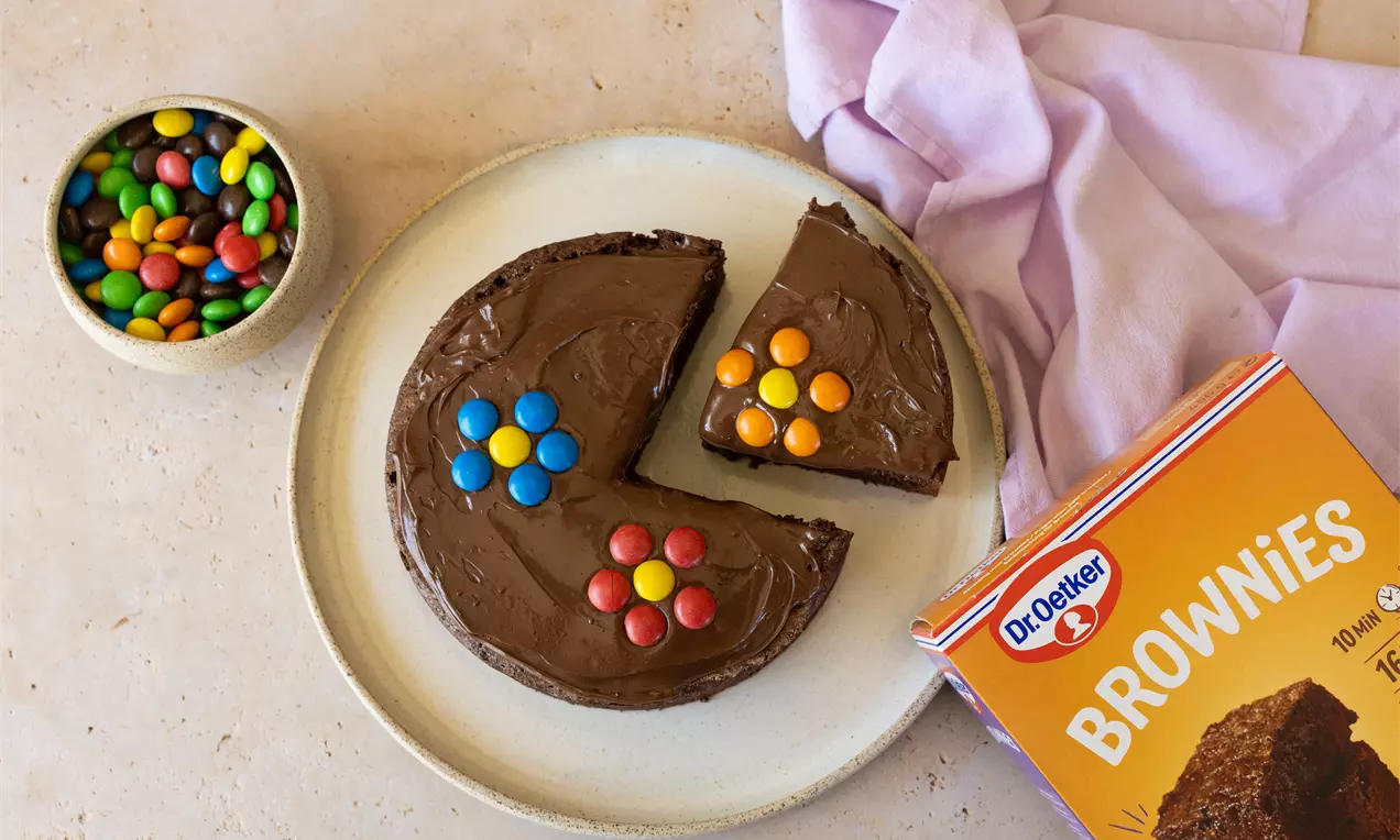 Ronde brownie met glanzende chocoladelaag, versierd met kleurrijke M&M-bloemen, op een wit bord met een schaaltje M&M’s en een paars servet