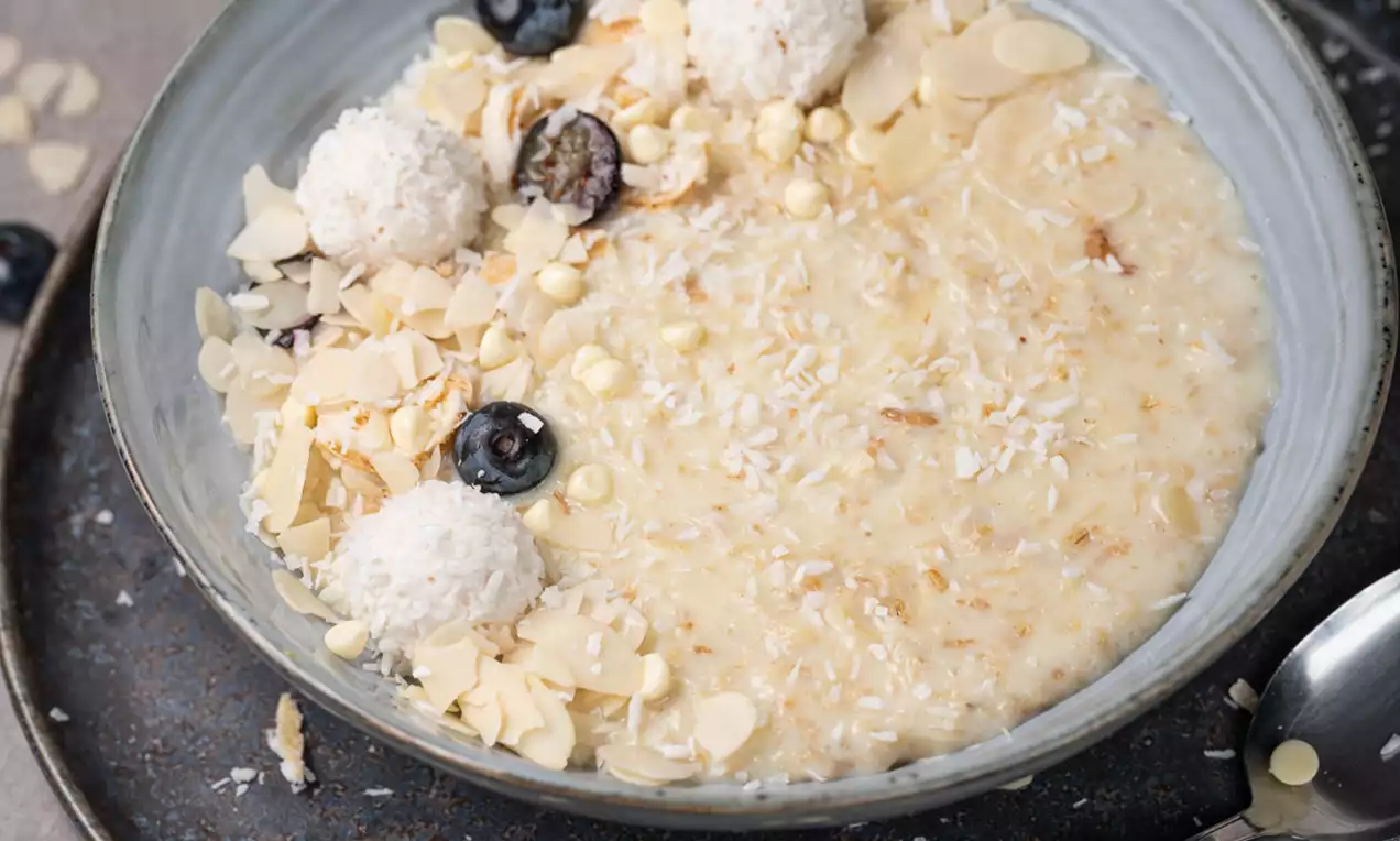 Svetla, kremasta kaša v beli skledi s celimi mandlji, kokosovimi kroglicami ter svežimi borovnicami na rustikalnem krožniku