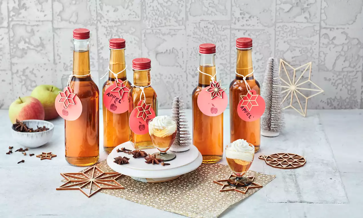 Mehrere schlanke Glasflaschen mit goldbraunem Likör und roten Verschlüssen, dekoriert mit roten Anhängern, auf hellem Untergrund mit weihnachtlichem Schmuck