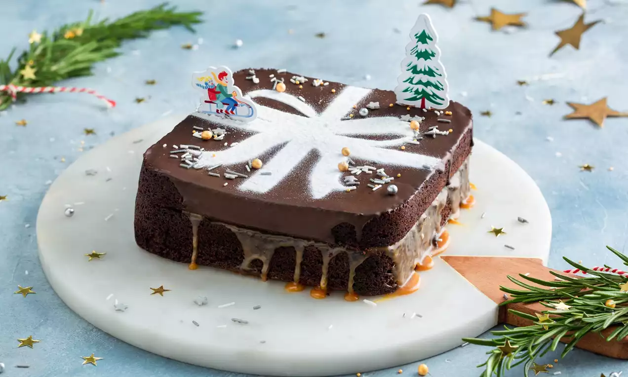 Vierkante kerstbrownie met glanzend bruine bovenkant, wit suikermotief, karamel aan de zijkanten en decoraties op een marmeren bord