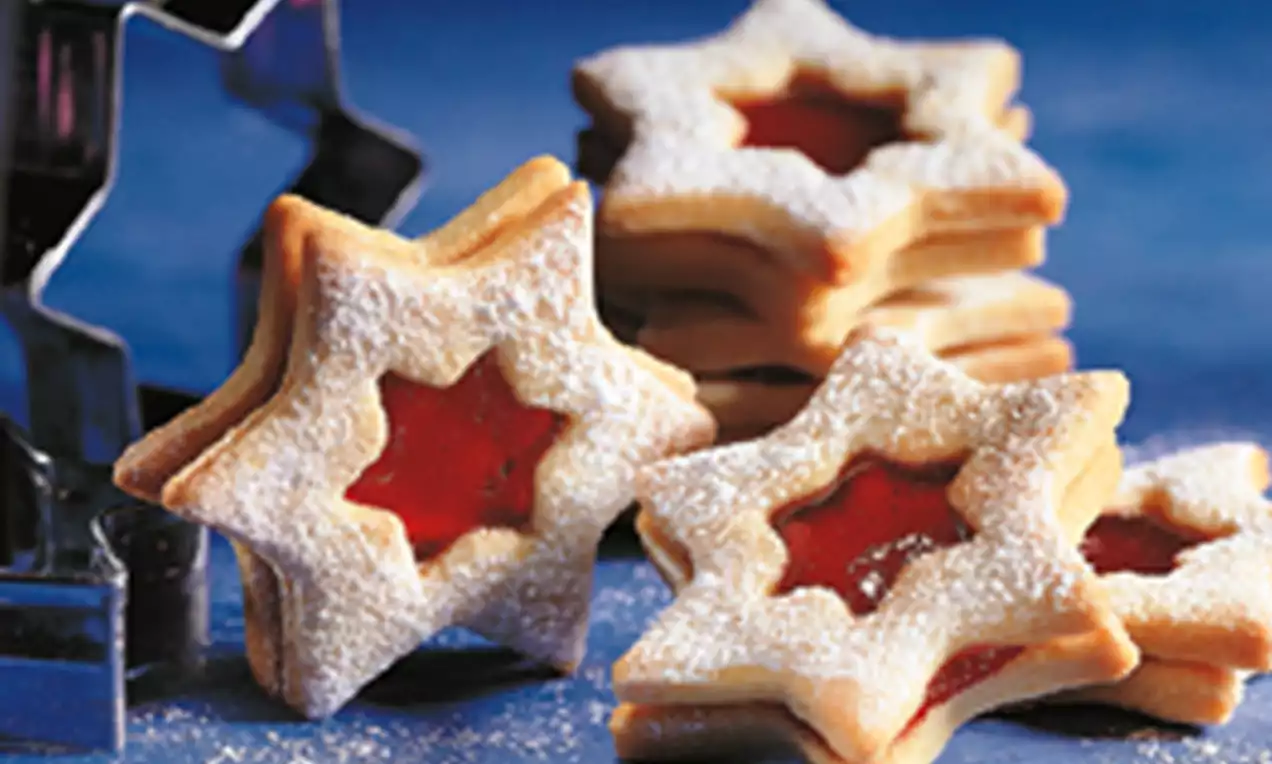 Stervormige boterkoekjes met een glanzende frambozenvulling, licht bestoven met witte poedersuiker op een blauwe ondergrond