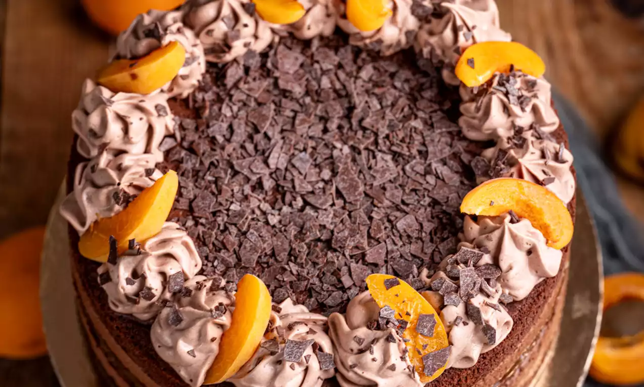 Čokoladna torta temne rjave barve z marelično marmelado, okrašena s čokoladnimi kosmiči, kremnimi rozetami in rezinami svežih marelic na leseni podlagi