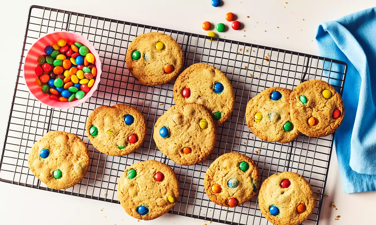 Golden brown cookies studded with vibrant candy pieces on a black cooling rack, with a pink bowl of candies and blue cloth nearby