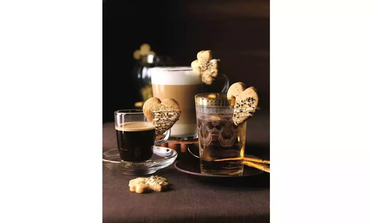 Sydämenmuotoiset vaaleat keksit, koristeina tummaa suklaaraitaa, aseteltuina lasien reunoille tummanruskealla pöydällä