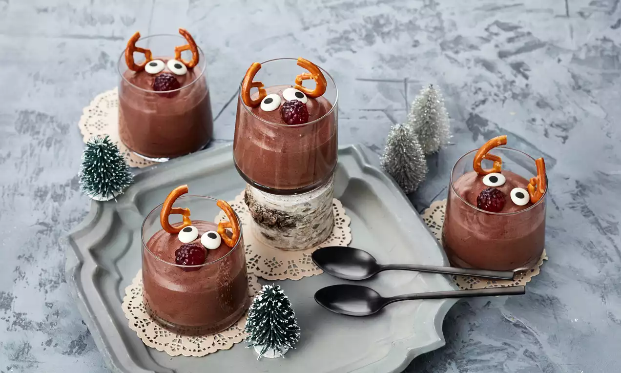 Vier Gläser mit schokoladenbraunem Dessert, verziert mit Brezeln als Geweih, Zuckeraugen und einer roten Beere, auf Tablett mit Mini-Tannenbäumen drapiert