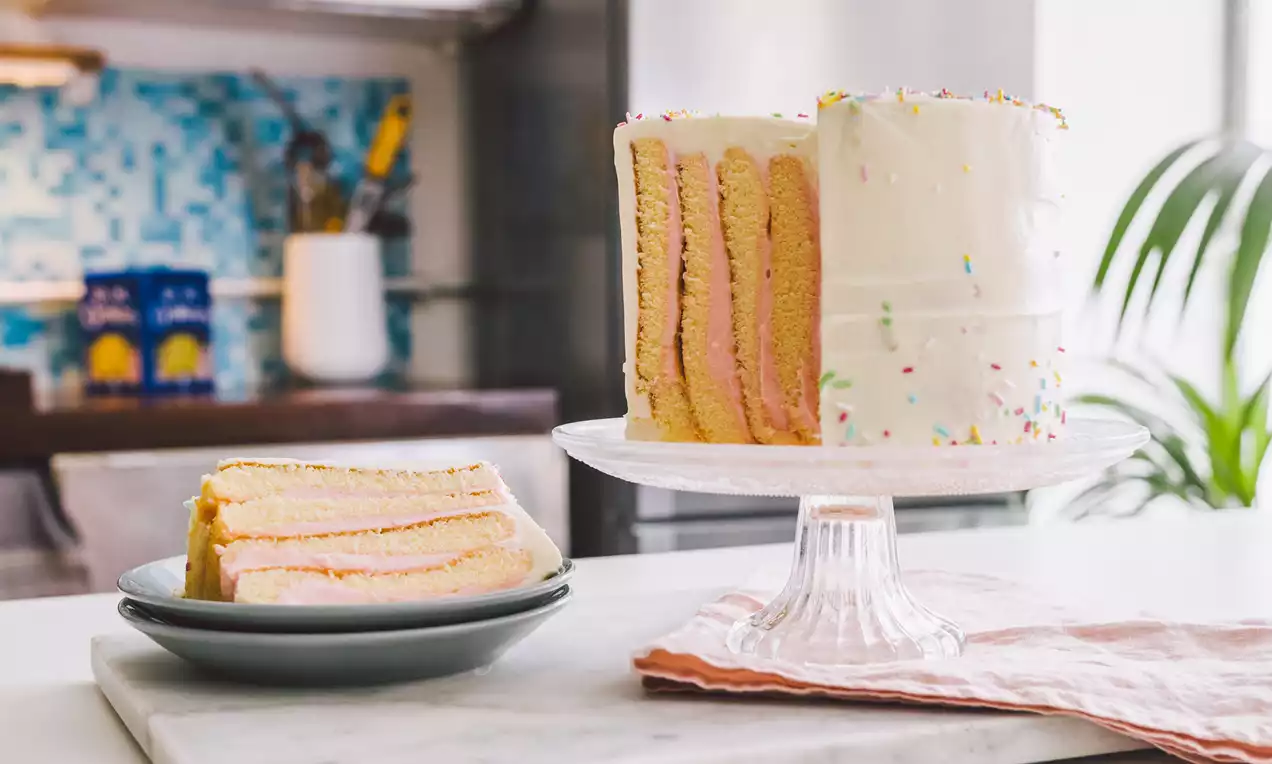 Tarta en capas verticales de bizcocho dorado y crema rosada, cubierta con glaseado blanco y detalles de confeti sobre un plato elevado