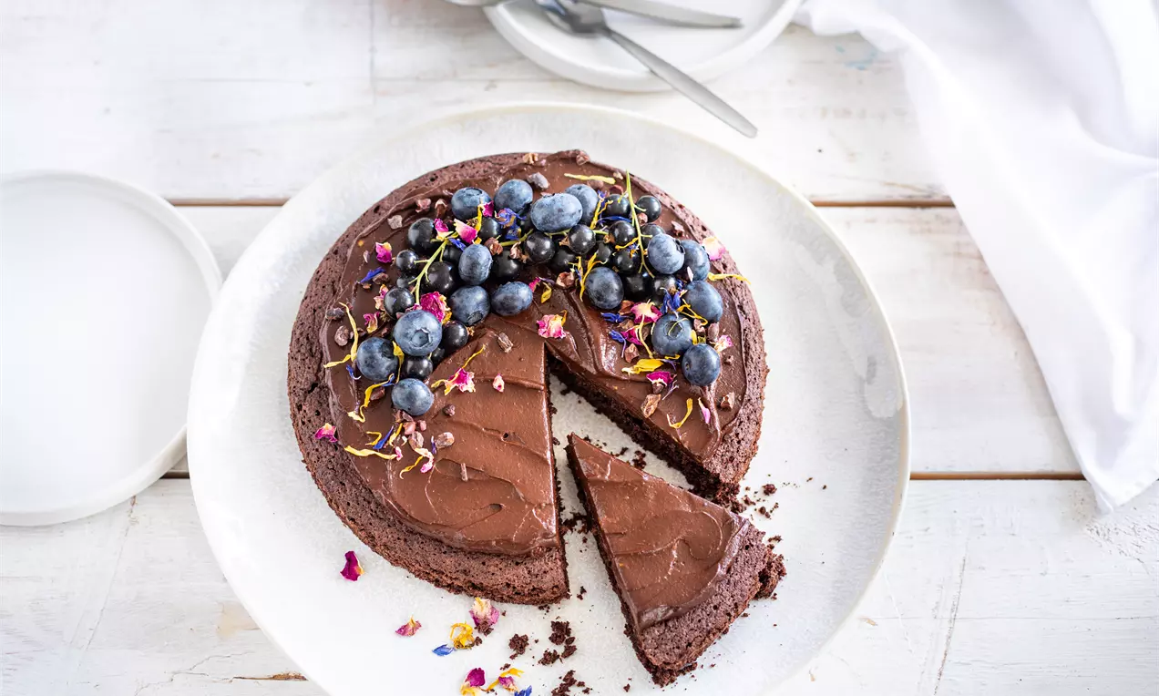 Кръгъл шоколадов сладкиш с гладка, тъмнокафява глазура, украсен с боровинки и ядливи цветя, върху бяла чиния на светъл фон