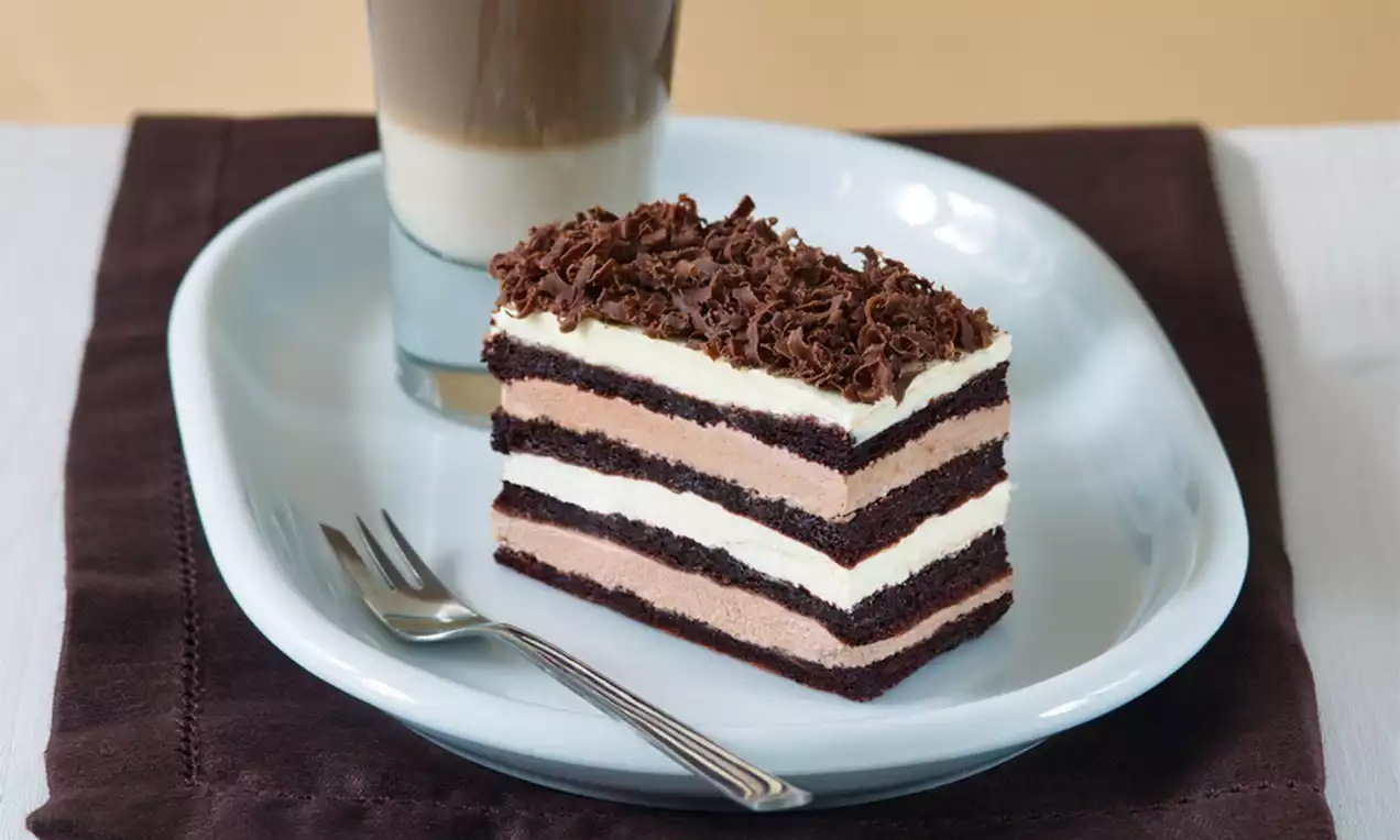 Vrstvený dezert s čokoládovými plátmi a krémovými výplňami, ozdobený nastrúhanou čokoládou na bledomodrom tanieri vedľa vidličky a latte