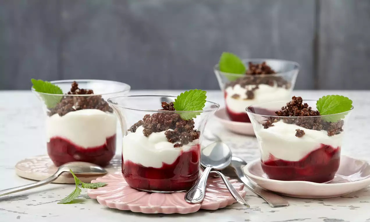 Gläser mit rotem Kompott, weißer Creme, knusprigen Schokobröseln und grünen Minzblättern auf rosa Tellern und heller Oberfläche
