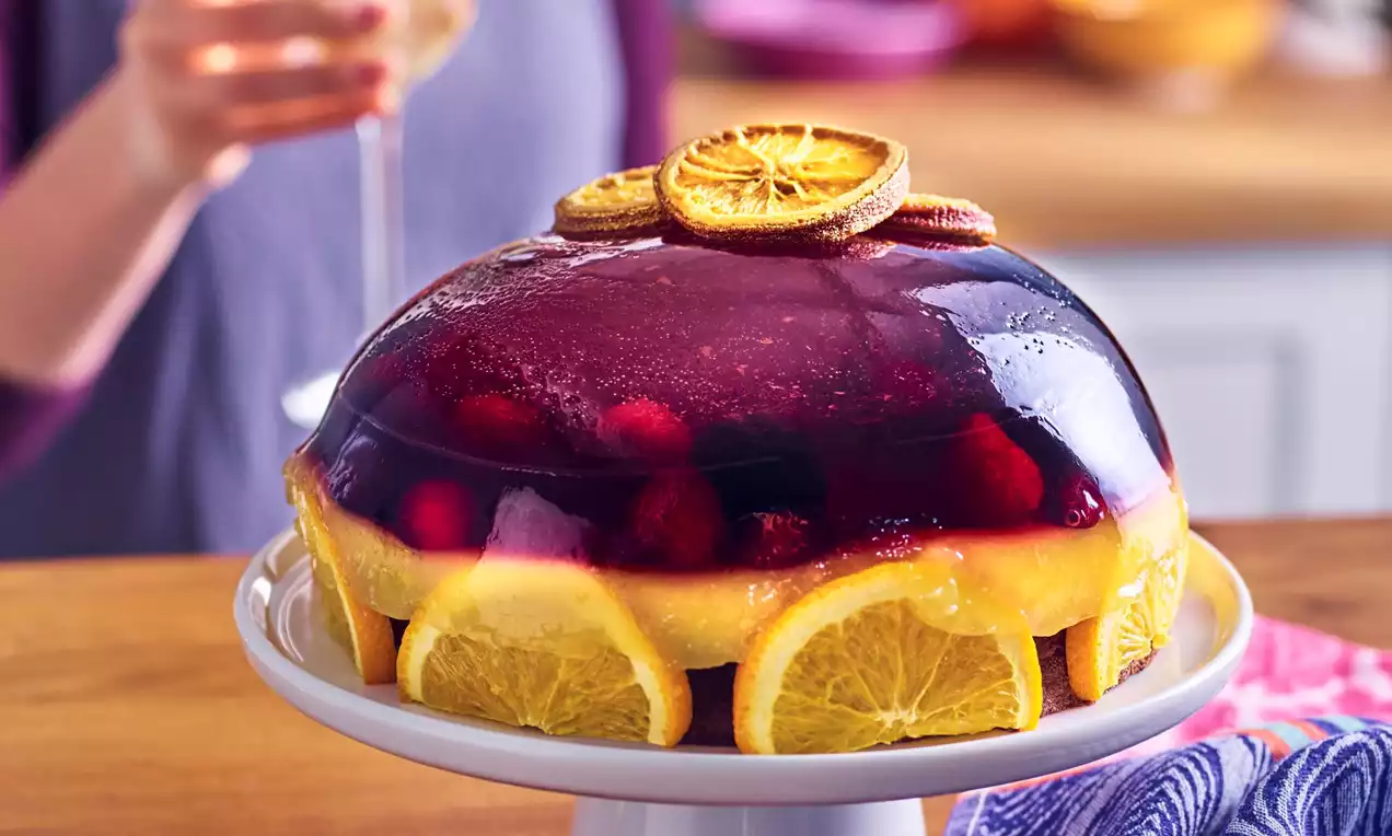 Layered jelly cake with a glossy deep red top, orange slices around the base, and dried orange garnishes on a white pedestal stand