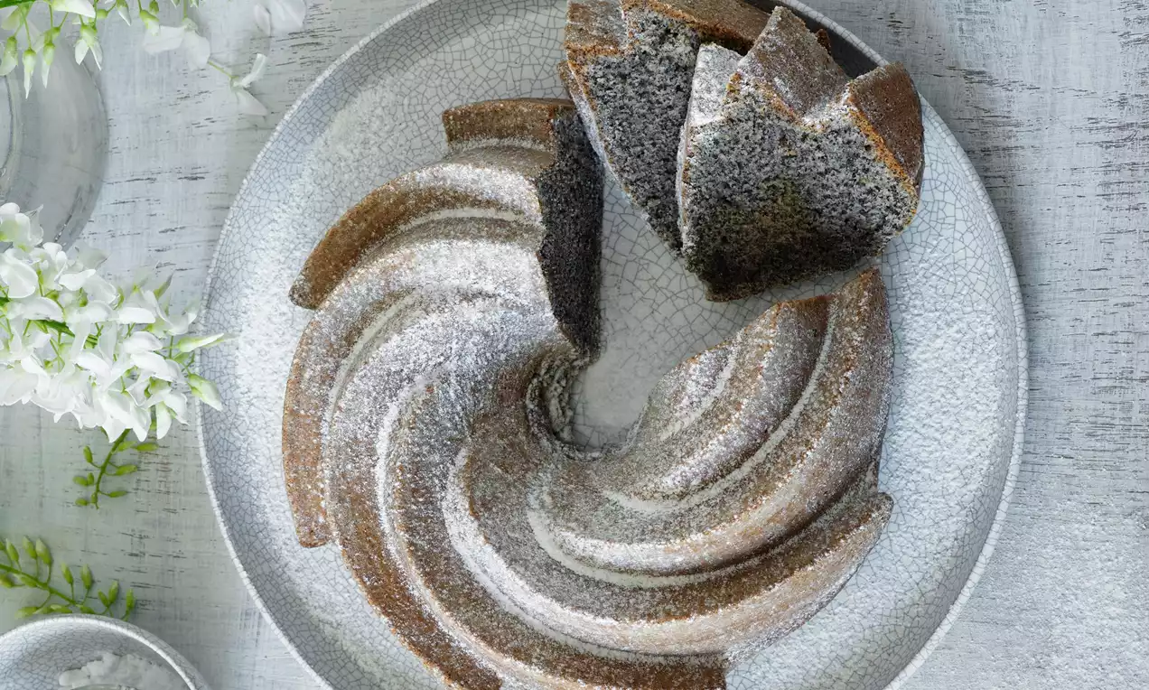 Tmavá maková bábovka ve spirálovém tvaru, jemně poprášená moučkovým cukrem, na světle šedém talíři vedle bílých květů a zelených lístků