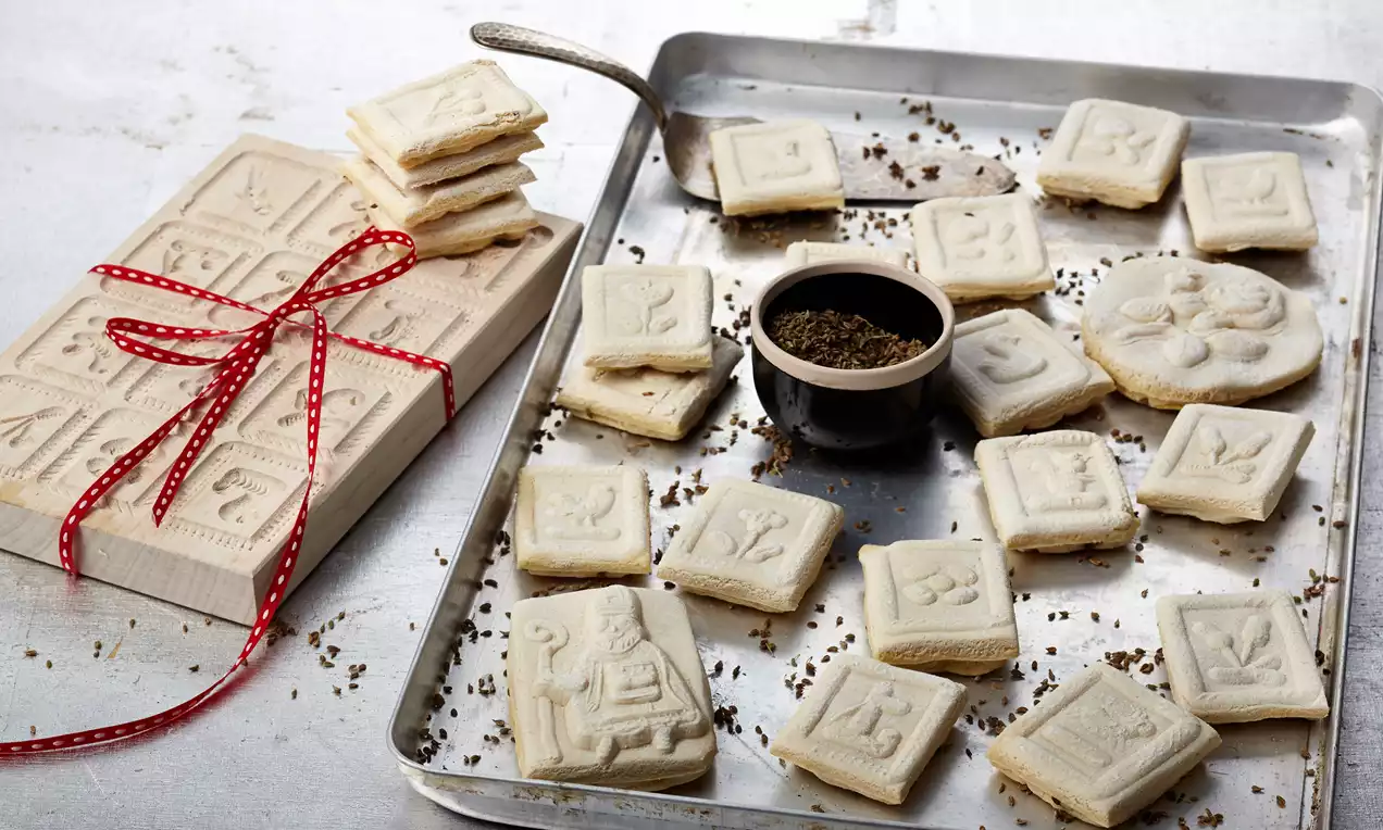 Rechteckige, helle Springerle mit geprägten Motiven auf silbernem Backblech neben Model aus Holz mit rotem Band und Becher mit Anis