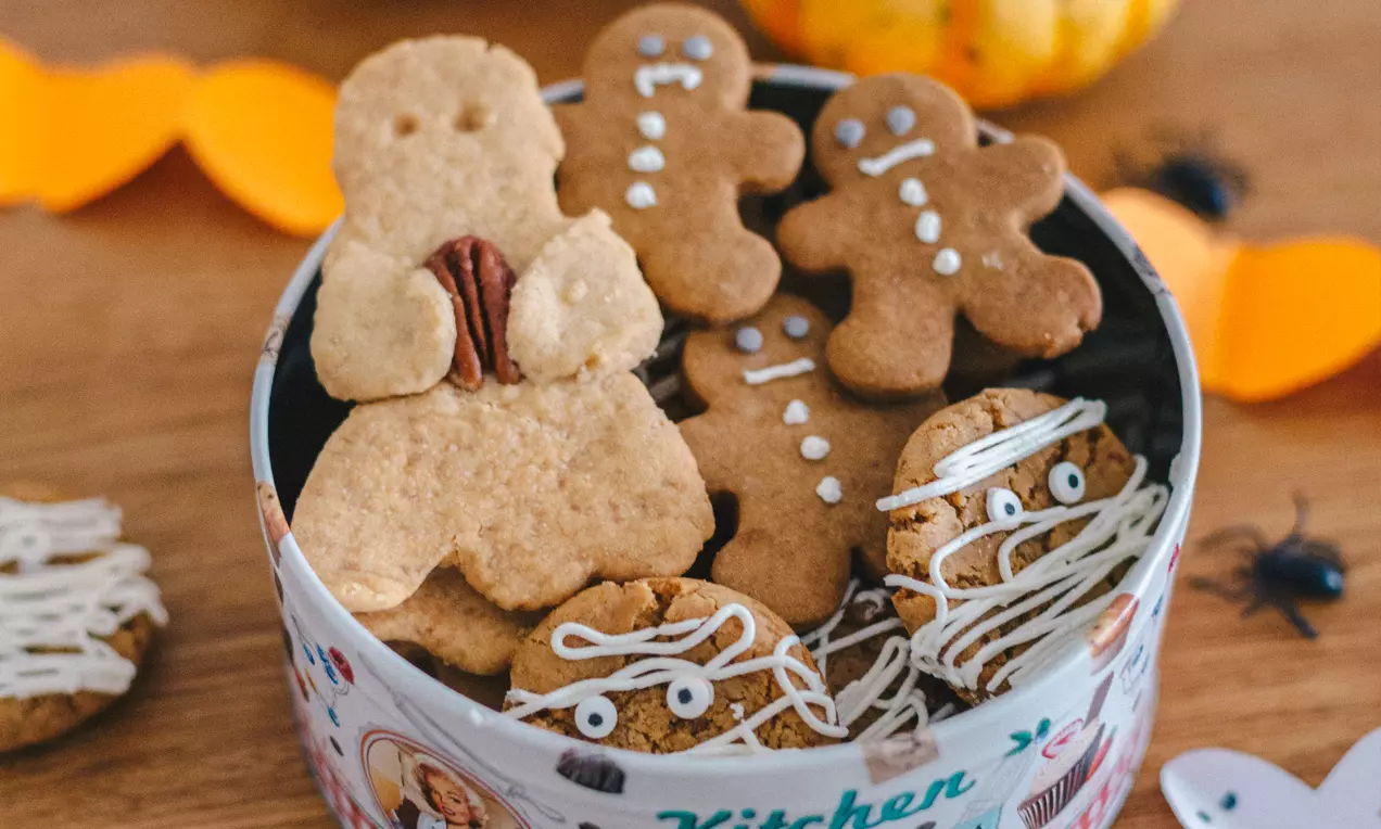 Карамелени сладки с човешки форми и шарки от бяла глазура, кръгли бисквити с шоколад и паяжини, наредени в метална кутия с шарени мотиви