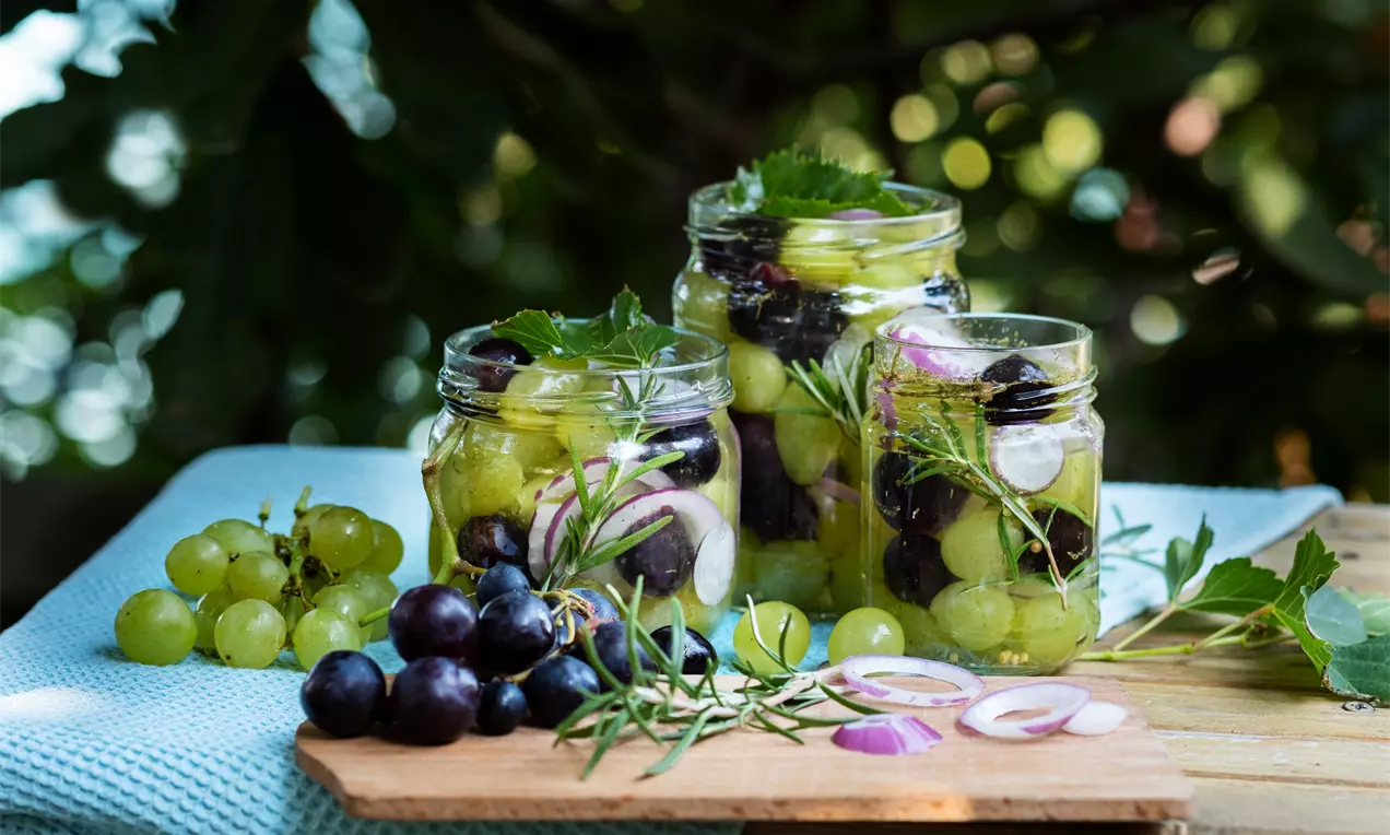 Туршия от зелени и тъмни гроздови зърна, розови шайби лук и стръкове розмарин в чисти буркани върху дървена поставка
