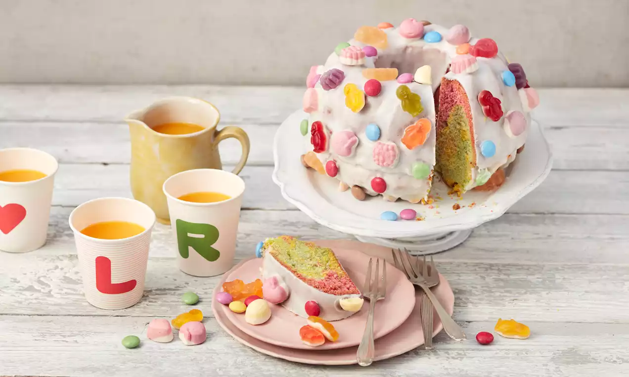 Runder Marmorkuchen mit Zuckerguss, bunt bestreut mit Süßigkeiten, auf einer Tortenplatte, daneben Teller mit Kuchenstück und Saftbecher