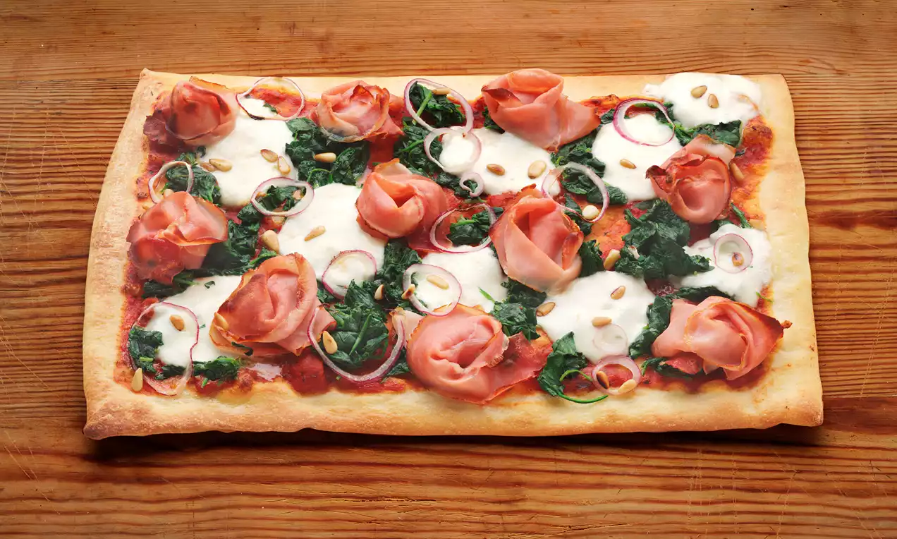 Rechthoekige pizza met een goudbruine korst, bedekt met groene spinazie, witte mozzarella, roze ham, uienschijfjes en pijnboompitten op houten tafel