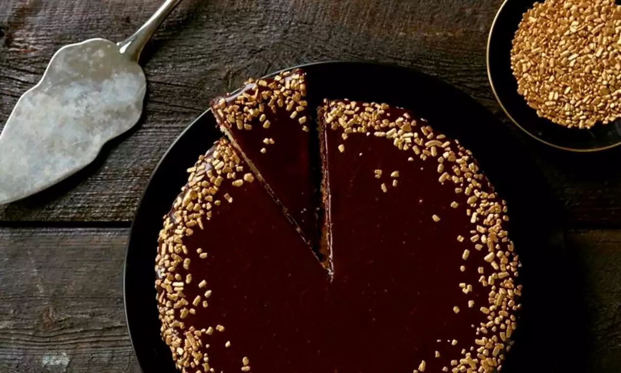 Konfektkake med mørk glasur og gullfarget strøssel langs kanten på et sort fat ved siden av en kakespade og en skål med strøssel