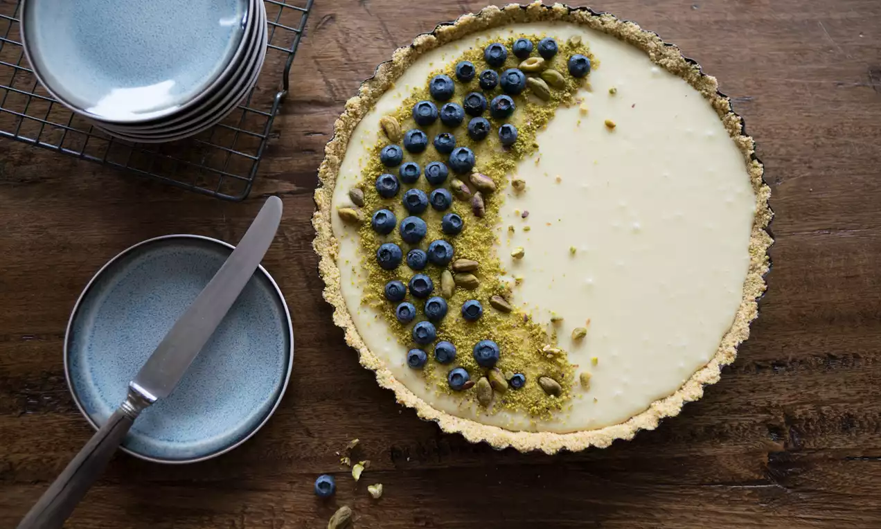 Witte chocolade ganache in een goudbruine taartkorst, half bedekt met blauwe bessen en pistachenoten, op een houten tafel met blauw servies