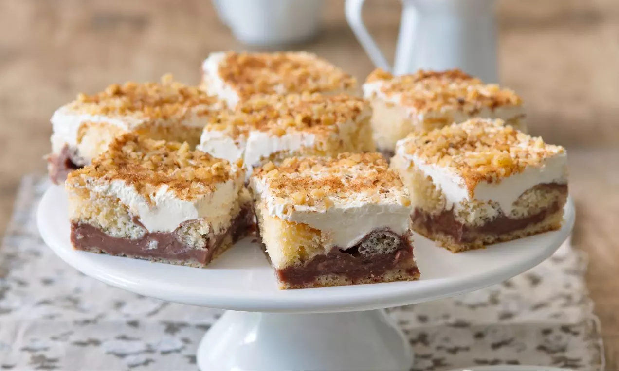 Štvorcové porcie vrstveného pudingového dezertu s čokoládovým spodkom, svetlým krémom a zlatistou strúhankou na bielom podnose